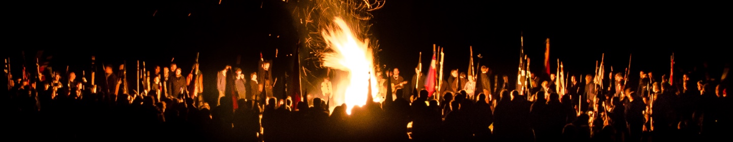 Ein großes Feuer in der Nacht. Viele Pfadfinderinnen und Pfadfinder stehen um es herum.